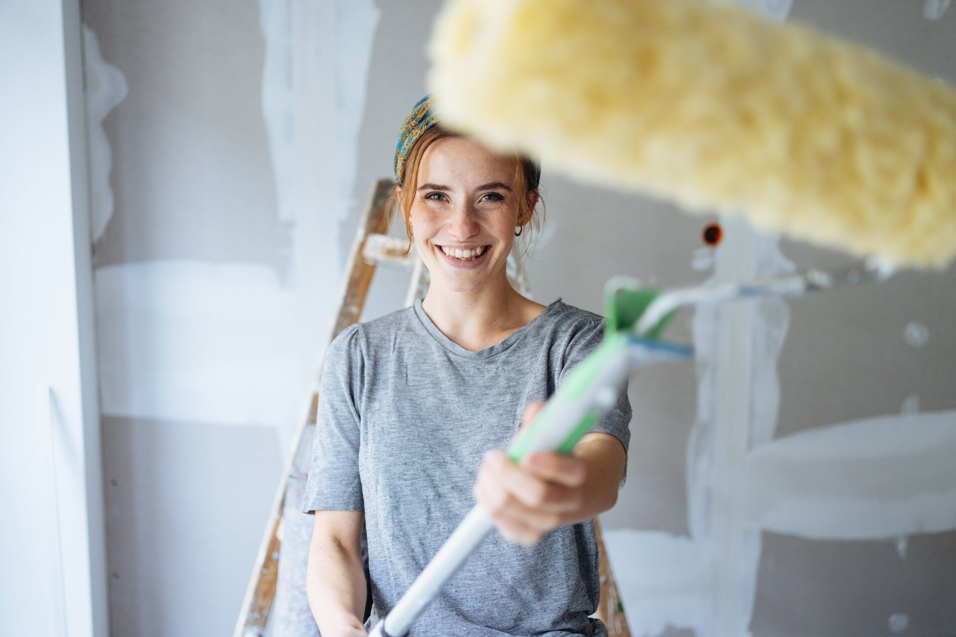 Lächelnde junge Frau, die einen Farbroller in die Kamera schwingt, während sie die Wände ihrer Wohnung streicht, bzw. DIY-Renovierungen durchführt.
